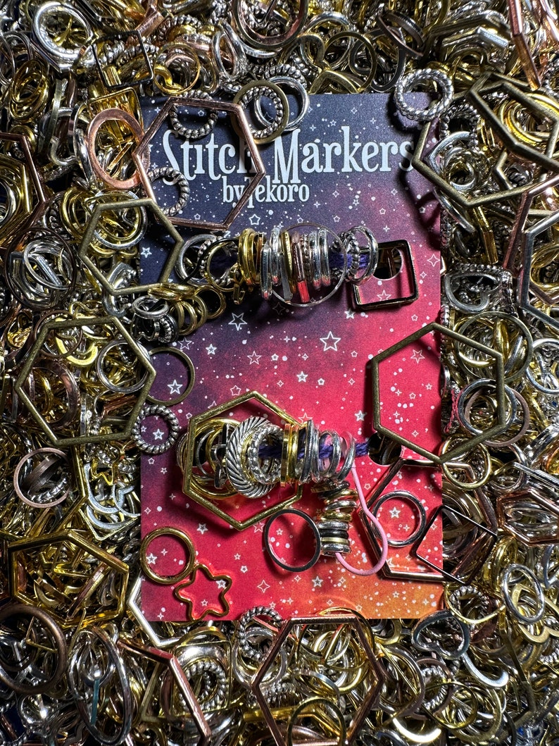May include: A close-up of a card with the text "Stitch Markers by Tekoro" on a red background with white stars. Two sets of stitch markers are displayed on the card, one set is silver and the other is gold. The stitch markers are arranged in a circle shape and are surrounded by a variety of metal rings and hoops.
