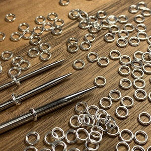 May include: A close-up of a large quantity of silver jump rings, along with two metal tools for jewelry making. The jump rings are scattered on a wooden surface.