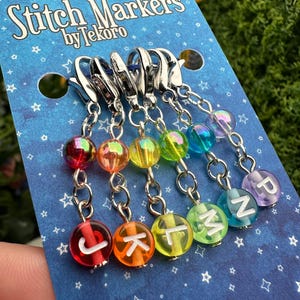 May include: A set of rainbow-colored stitch markers with silver clasps. Each marker has a clear, round bead with a letter inside. The card reads "Stitch Markers by Tekoro" in white text on a blue background with star patterns.