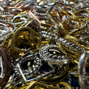 May include: A close-up of a pile of various metal charms and pendants in gold, silver, and copper tones. The charms are in different shapes, including circles, hearts, and squares. Some of the charms have intricate designs, while others are plain.
