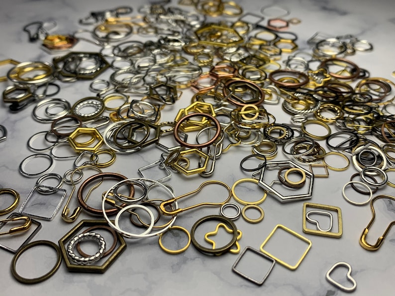 May include: A large assortment of metal jump rings, charms, and other jewelry findings in gold, silver, and bronze tones. The findings are scattered on a white surface.