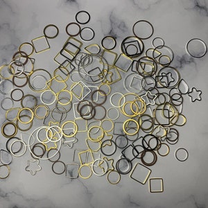 May include: A collection of gold, silver, and black metal rings and squares of various sizes. The shapes are scattered on a white marble surface.