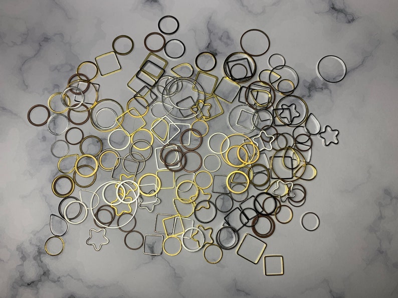May include: A collection of gold, silver, and black metal rings and squares of various sizes. The shapes are scattered on a white marble surface.