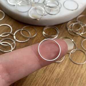 Puede incluir: Primer plano de un anillo de salto plateado sostenido entre un dedo y el pulgar. El anillo es peque&ntilde;o y circular, y hay muchos otros anillos similares esparcidos en el fondo.