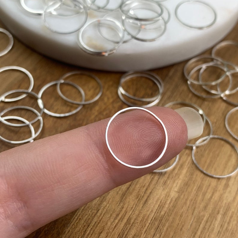 Puede incluir: Primer plano de un anillo de salto plateado sostenido entre un dedo y el pulgar. El anillo es peque&ntilde;o y circular, y hay muchos otros anillos similares esparcidos en el fondo.