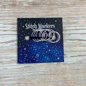 Puede incluir: Un paquete de marcadores de punto de plata con un fondo azul con estrellas blancas. El paquete dice "Stitch Markers by Tekoro".