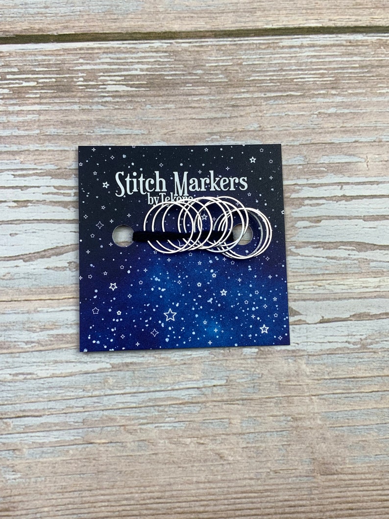 Puede incluir: Un paquete de marcadores de punto de plata con un fondo azul con estrellas blancas. El paquete dice "Stitch Markers by Tekoro".