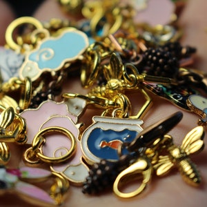 May include: A collection of colorful enamel charms featuring a variety of designs, including a bee, a fishbowl, a cloud, a flower, and a bunny. The charms are made of metal and have a gold finish.