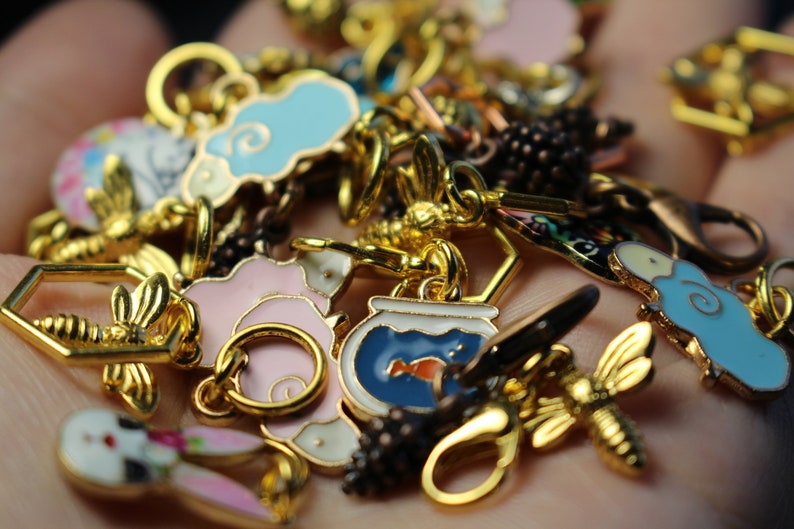 May include: A collection of colorful enamel charms featuring a variety of designs, including a bee, a fishbowl, a cloud, a flower, and a bunny. The charms are made of metal and have a gold finish.