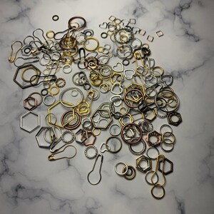 May include: A large collection of assorted gold, silver, and bronze jump rings, key rings, and other metal findings. The rings are scattered on a white marble surface.