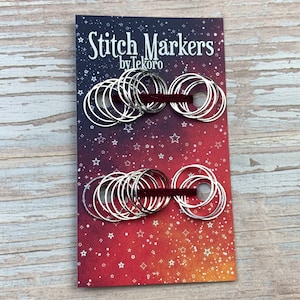 Puede incluir: Un paquete de marcadores de punto de plata con una cinta roja. El paquete tiene un fondo rojo, naranja y amarillo con estrellas blancas. El texto en el paquete dice "Stitch Markers by Tekoro".