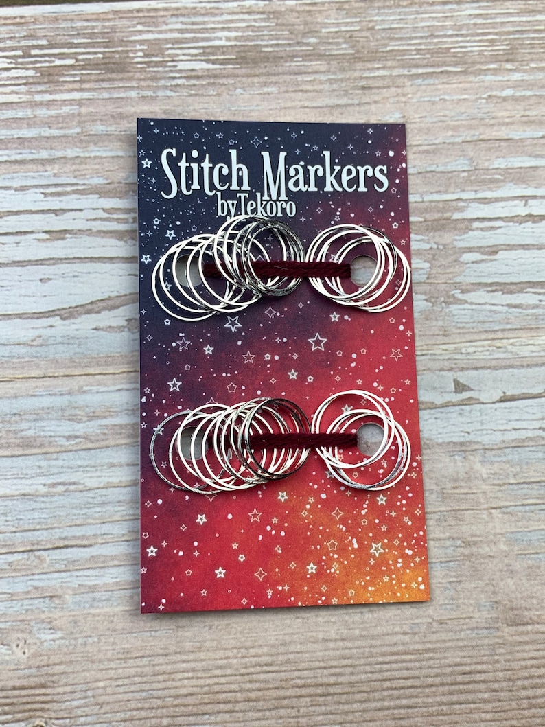 Puede incluir: Un paquete de marcadores de punto de plata con una cinta roja. El paquete tiene un fondo rojo, naranja y amarillo con estrellas blancas. El texto en el paquete dice "Stitch Markers by Tekoro".