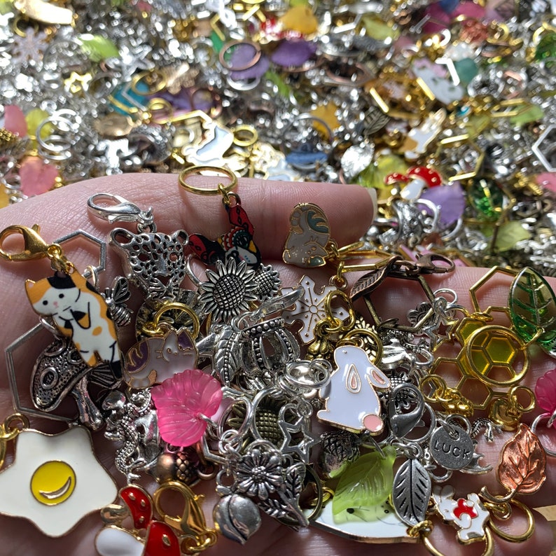 May include: A collection of charms featuring various designs, including a cat, a rabbit, a sunflower, a crown, a snowflake, a honeycomb, and a leaf. The charms are made of metal and have a silver, gold, and white color scheme.