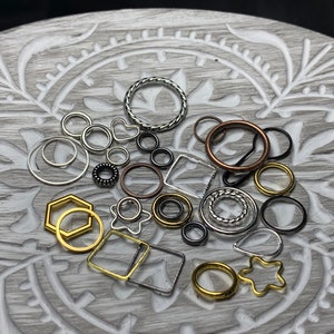 May include: Assortment of silver, gold, and copper jump rings in various shapes and sizes. The jump rings are arranged on a gray and white patterned surface.