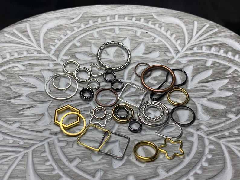May include: Assortment of silver, gold, and copper jump rings in various shapes and sizes. The jump rings are arranged on a gray and white patterned surface.