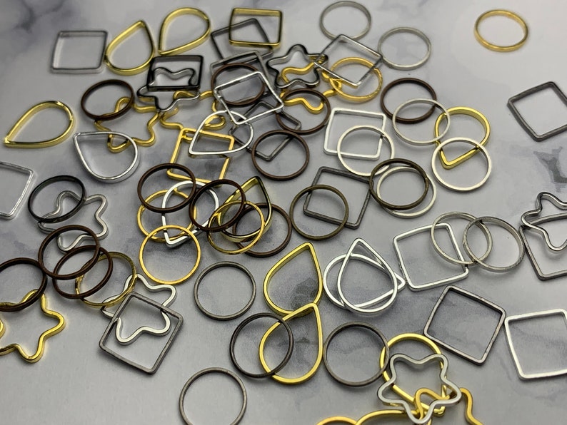 May include: Assortment of metal jump rings in various shapes and sizes, including circles, squares, teardrops, and stars. The jump rings are in gold, silver, and bronze tones.