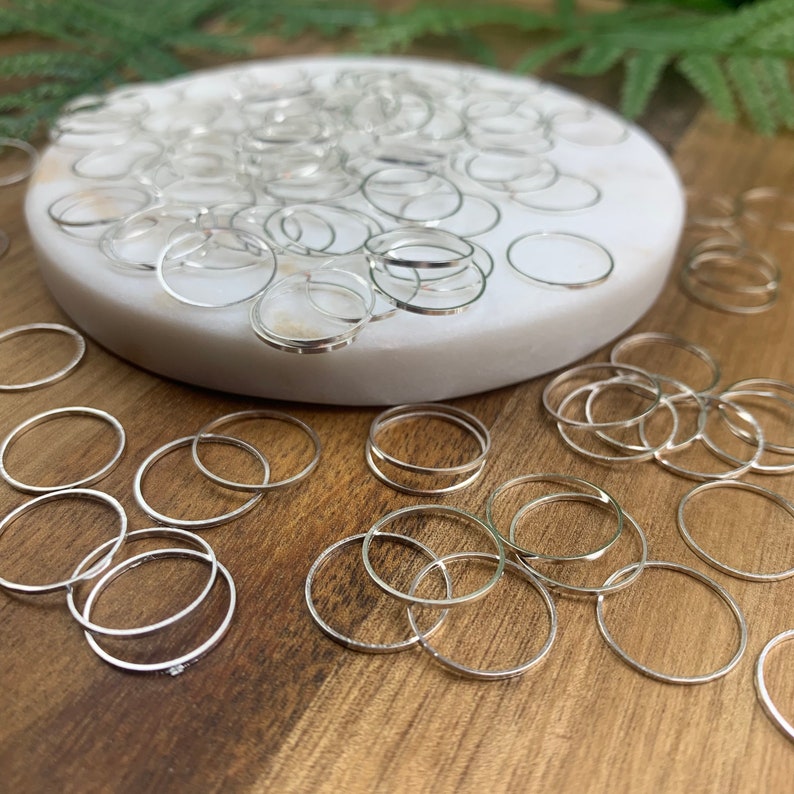 Puede incluir: Una gran cantidad de anillos de salto plateados, algunos est&aacute;n apilados sobre una superficie blanca, otros est&aacute;n dispersos sobre una superficie de madera.