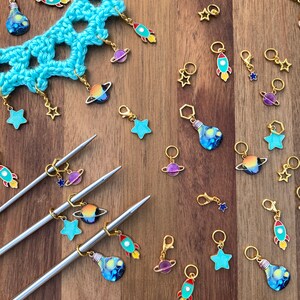 May include: A collection of colorful stitch markers with various designs, including planets, rockets, light bulbs, and stars. The markers are made of metal and have a gold finish. They are arranged on a wooden surface with two knitting needles.