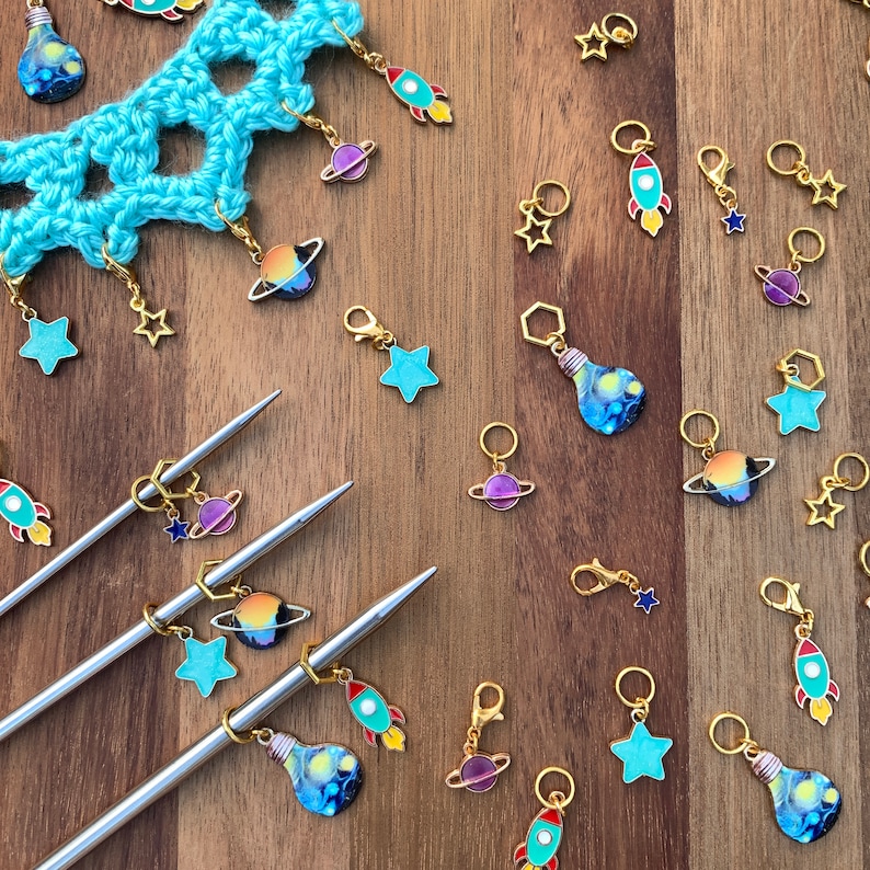 May include: A collection of colorful stitch markers with various designs, including planets, rockets, light bulbs, and stars. The markers are made of metal and have a gold finish. They are arranged on a wooden surface with two knitting needles.