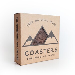 May include: A brown cardboard box with the text "THE MOUNTAINS ARE CALLING" on the side. The front of the box has a graphic of mountains with the text "100% NATURAL WOOD" and "COASTERS FOR MOUNTAIN PEOPLE".