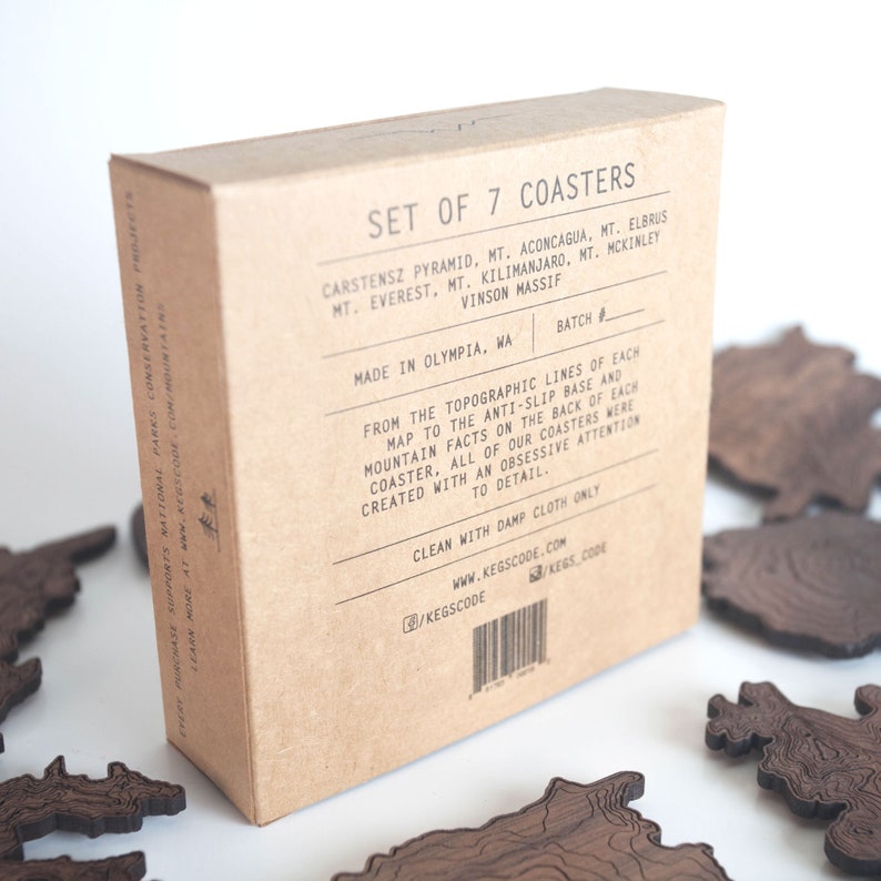 May include: A set of seven brown wooden coasters featuring topographic maps of seven mountains: Carstensz Pyramid, Aconcagua, Elbrus, Everest, Kilimanjaro, McKinley, and Vinson Massif. The coasters are made in Olympia, Washington. The box features a batch number and a website address.