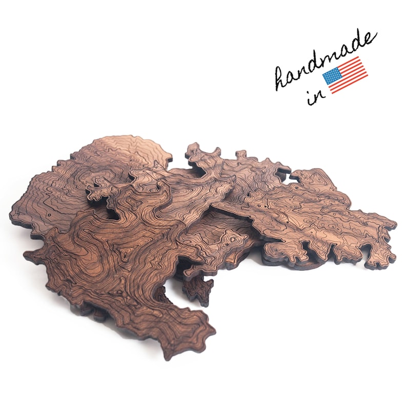 May include: A wooden map of a mountainous region, with intricate lines representing elevation contours. The map is made of dark brown wood and has a natural, rustic look. The text "handmade in" is written in black ink, with a small American flag next to it.