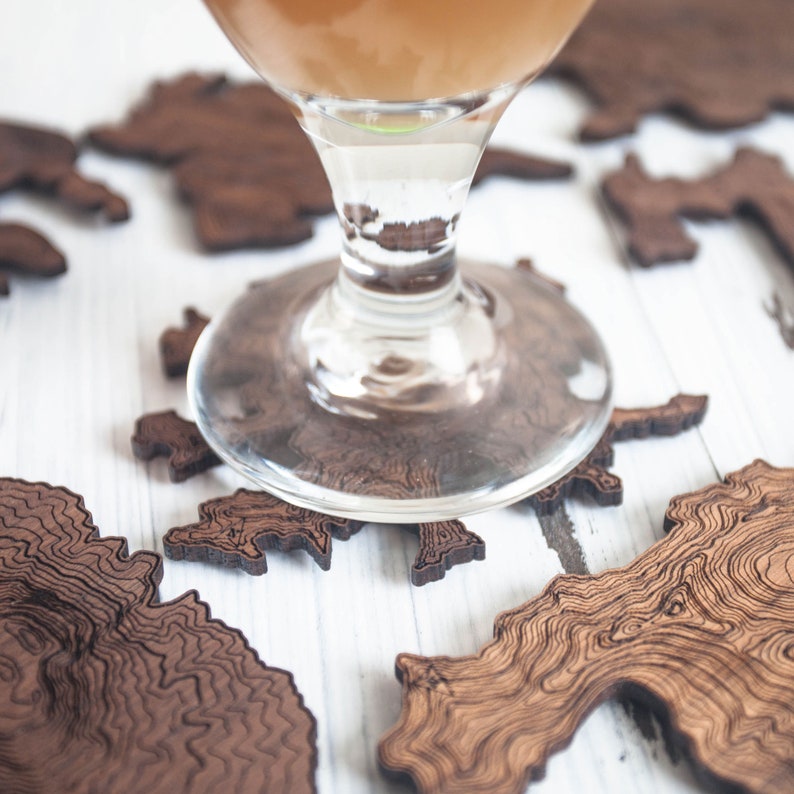 May include: A glass of amber-colored liquid sits on a set of wooden coasters. The coasters are shaped like islands and have a detailed wood grain pattern.