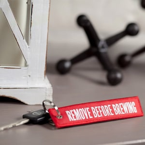 Könnte beinhalten: Roter Stoffschlüsselanhänger mit weißem Text "REMOVE BEFORE BREWING".
