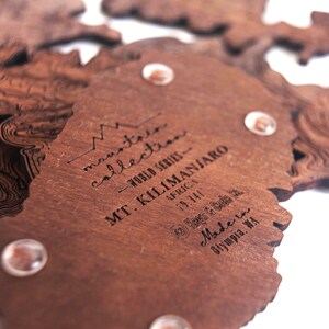 May include: A wooden map of Mount Kilimanjaro, part of the "mountain collection - world series" with the text "MT. KILIMANJARO - AFRICA - 19,341" and "Made in Olympia, WA".