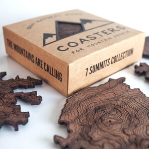 May include: A set of wooden coasters with a topographic map design. The coasters are in a brown cardboard box with the text "COASTERS FOR MOUNTAIN PEOPLE" and "7 SUMMITS COLLECTION". The box also has the text "THE MOUNTAINS ARE CALLING" on the side.