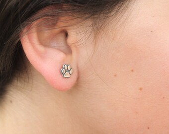 May include: A close-up of a person's ear with a small silver paw print stud earring.
