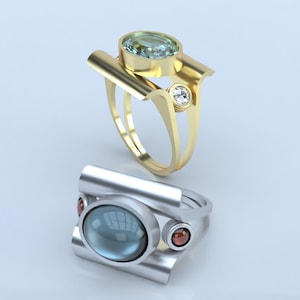 May include: Two rings, one gold and one silver, each featuring a large, oval gemstone. The gold ring has a light green stone and a small clear stone. The silver ring has a blue-green stone and two small red stones. Modern jewelry.