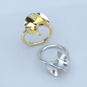 Może przedstawiać: Dwa pierścionki, jeden złoty i jeden srebrny, każdy o unikalnym, abstrakcyjnym wzorze. Pierścionki mają płynny, przypominający płatek kształt na górze obrączki. Złoty pierścionek ma jasne, błyszczące wykończenie, a srebrny matowe.