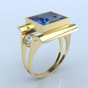 May include: A gold ring featuring a large, square, dark blue gemstone set in a rectangular bezel. A smaller, round, clear gemstone is set on the side. The ring has a unique, modern design with a double band.