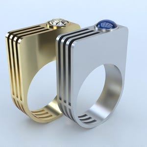 May include: Two modern rings, one gold-toned with a clear gemstone and the other silver-toned with a blue gemstone. Both rings have a rectangular top with three black stripes and a rounded band.