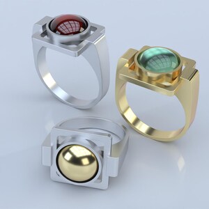 May include: Three rings with geometric designs. One ring is silver with a red gemstone, another is gold with a green gemstone, and the third is silver with a gold sphere. The rings have a modern, minimalist style.