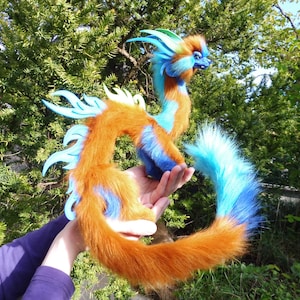 Può includere: Un giocattolo di drago di peluche con accenti arancioni, blu e verde acqua. Il drago ha una lunga coda soffice e pinne decorative. Il giocattolo è tenuto contro uno sfondo di fogliame verde.