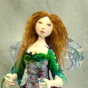 May include: A handmade doll with red hair wearing a green and purple dress with a green and purple bug. The doll is holding green strings with beads.