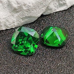 May include: Two faceted, emerald-green gemstones. One is square-shaped, the other is a rounded square. The gems are displayed on a dark, textured surface with a white, rough-textured rock in the background.