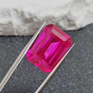 May include: A vibrant, rectangular-cut, hot pink gemstone is held by silver tweezers. The gem has a faceted surface that reflects light, showcasing its clarity and color. The background is a mix of gray and white.