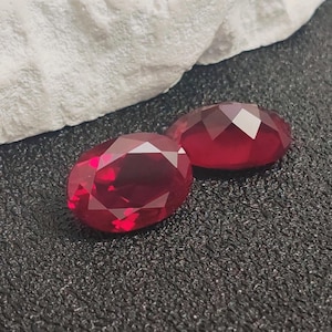 Pode incluir: Duas pedras preciosas vermelhas de formato oval, lapidadas com facetas e corte brilhante. As pedras exibem um tom carmesim profundo e saturado, posicionadas sobre uma superfície escura e texturizada perto de um material branco, rochoso e irregular.