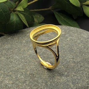 May include: A gold ring with an oval top and a triangular side design, set on a grey stone. Green leaves are visible in the background. The ring is likely a jewellery finding. The ring's design is modern and elegant.