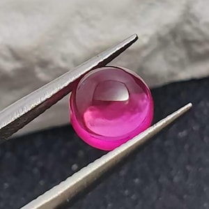 Può includere: Una gemma sferica, lucidata, di colore magenta, è tenuta da una pinzetta d'argento. La gemma ha una superficie liscia e riflettente, e la pinzetta è posizionata su uno sfondo neutro.