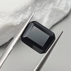 Puede incluir: Una gema negra rectangular sostenida por unas pinzas plateadas. La gema tiene una superficie pulida y está colocada sobre un fondo blanco texturizado. Las pinzas están enfocadas, resaltando la claridad de la gema.