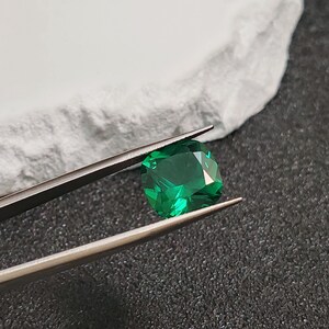 May include: A faceted, emerald-green gemstone is held by silver tweezers against a dark gray surface. The gem has a cushion cut, reflecting light. A white, textured surface is in the background.