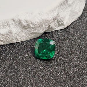 May include: A vibrant, emerald-cut green gemstone with a faceted surface. The gem is displayed on a dark, textured surface next to a white, rough-hewn stone.