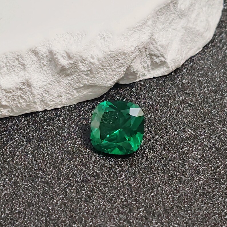 May include: A vibrant, emerald-cut green gemstone with a faceted surface. The gem is displayed on a dark, textured surface next to a white, rough-hewn stone.