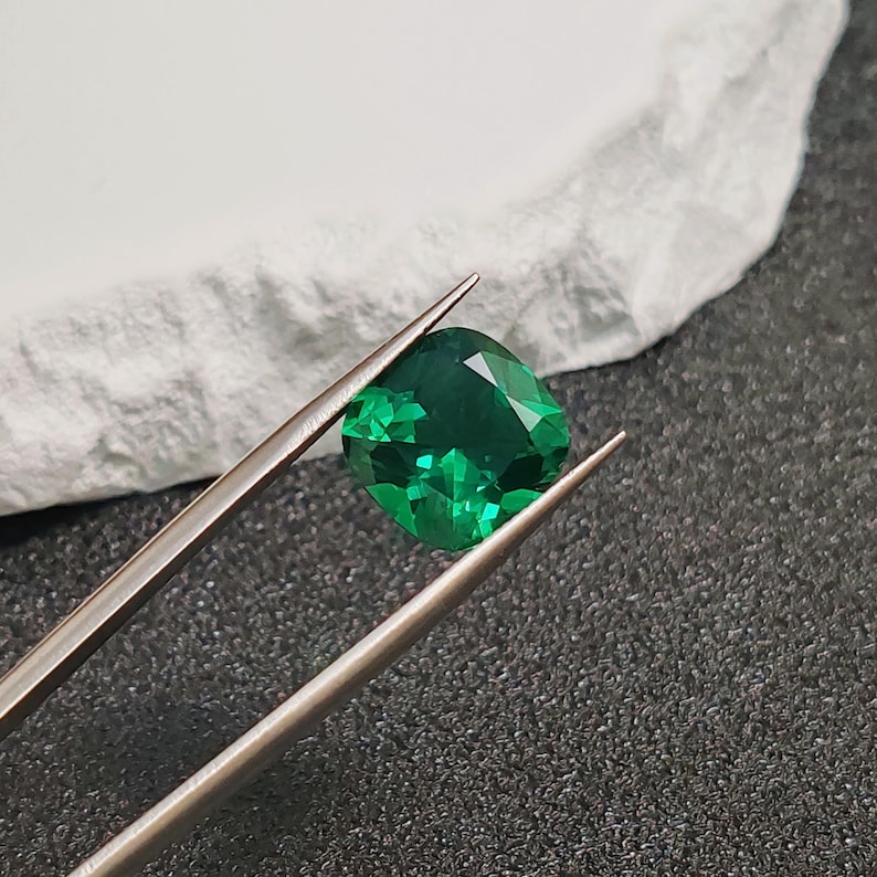May include: A vibrant, emerald-green gemstone, held by silver tweezers. The cushion-cut gem displays facets that catch the light, showcasing its clarity and brilliance. The background is a textured, dark surface with a white element.