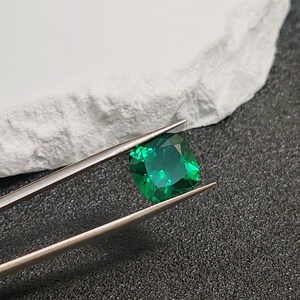 May include: A vibrant, emerald-cut green gemstone held by silver tweezers. The gem is a deep, saturated green with visible facets. The background includes a white, textured surface and a dark, speckled surface.