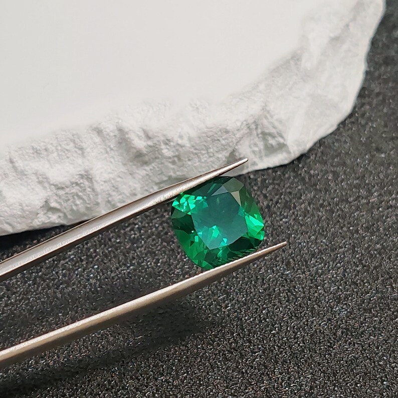 May include: A vibrant, emerald-cut green gemstone held by silver tweezers. The gem is a deep, saturated green with visible facets. The background includes a white, textured surface and a dark, speckled surface.